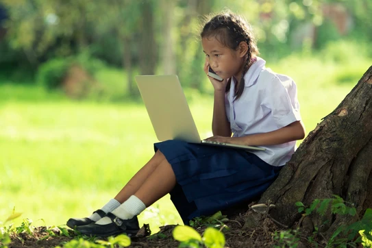 Girl sitting at the base of a tree on a laptop talking on the phone.