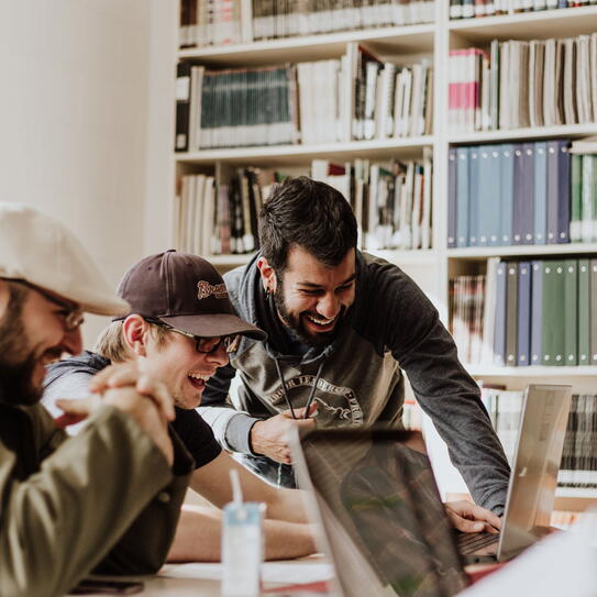 A group of people sitting around a desk with a laptops laughing.